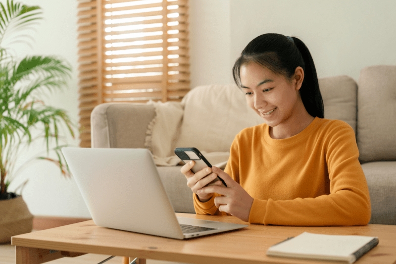 Young woman reviewing no credit check personal loan options on her smartphone while using a laptop at home